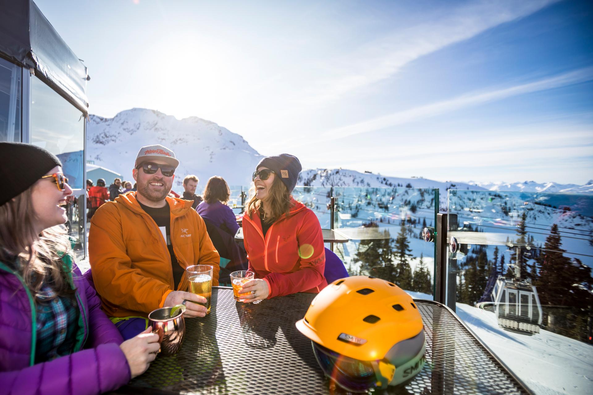 A group of friends enjoys drinks at a sunny mountain terrace with snow-covered slopes and ski lifts in the background.