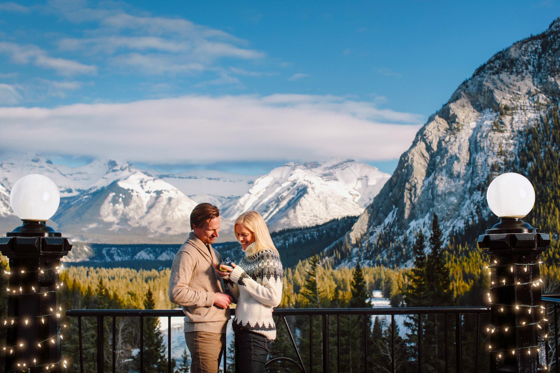 A smiling man and woman standing at a viewpoint with the snowy Rocky Mountains in the background; the woman is holding a small gift wrapped box.