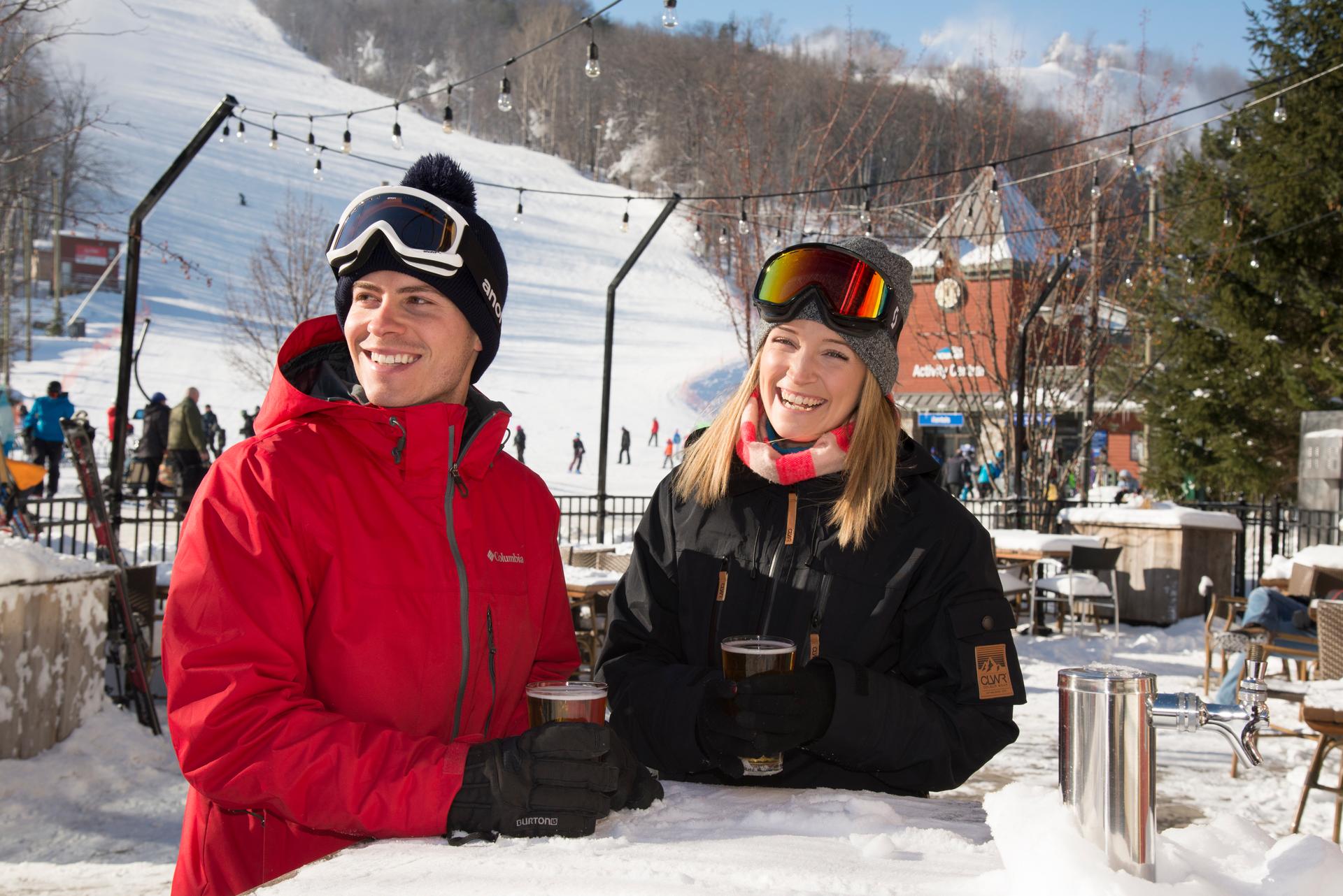 A man and a woman in ski gear smiling and drinking beer outdoors at the Blue Mountain ski area.