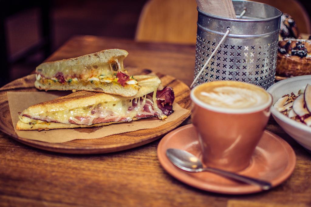 A close up photo of a toasted cheese and meat sandwich on a table next to a latte in a mug.