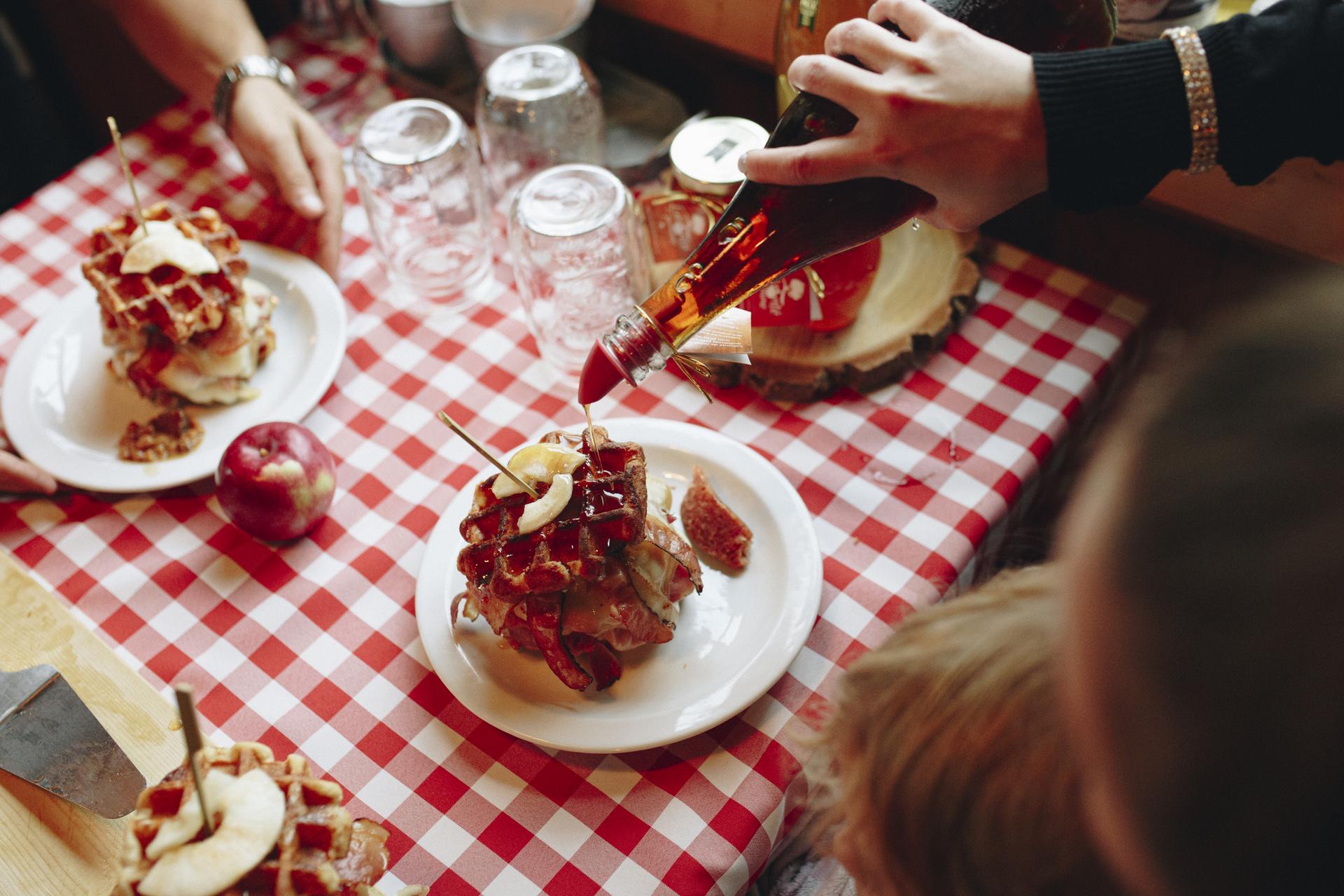 Maple syrup being drizzled on a stack of waffles and bacon