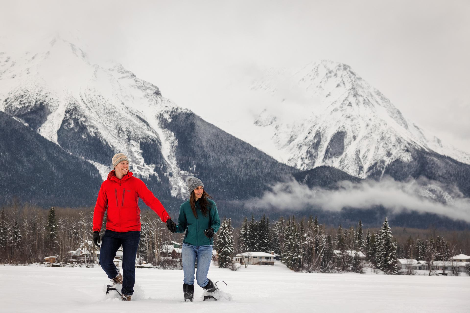 Two people snowshoeing across a flat expanse with tall snow-capped mountains in the background.