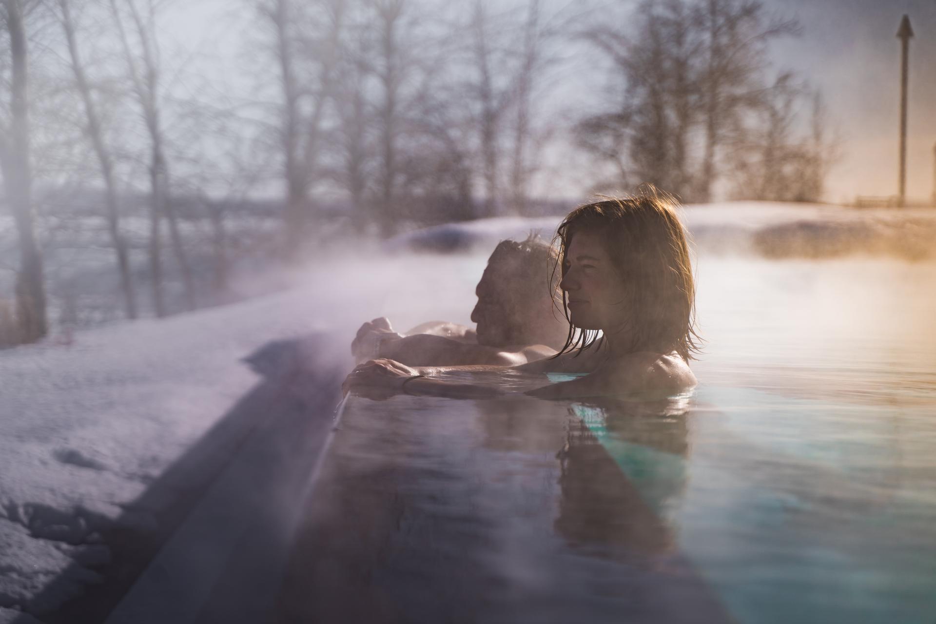 Two people leaning on the edge of an outdoor hot tub, looking out at the snowy view.