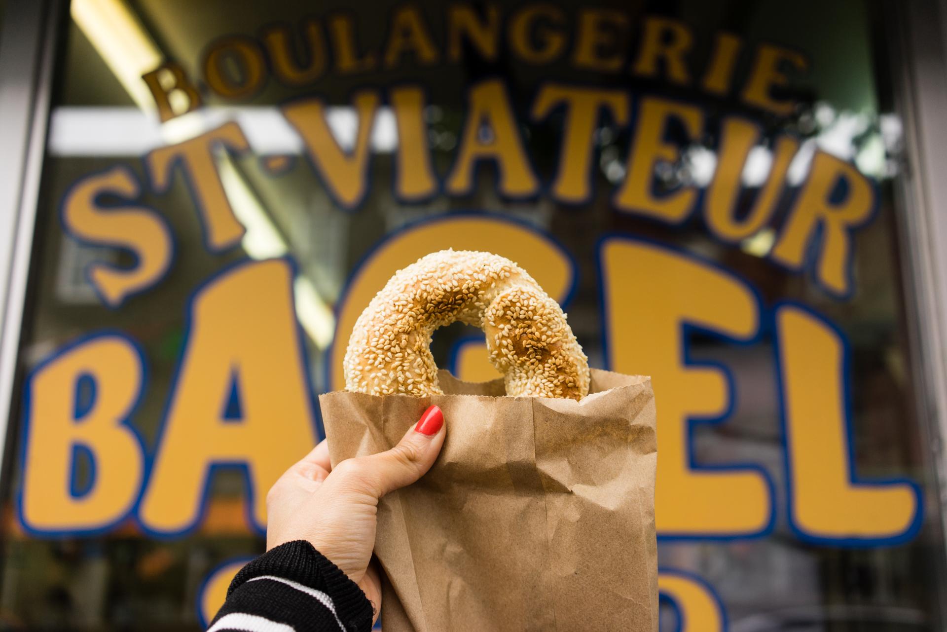 A hand with red fingernail polish holding up a paper bag with a sesame seed bagel sticking out halfway in front of the window for St-Viateur Bagel in Montreal.