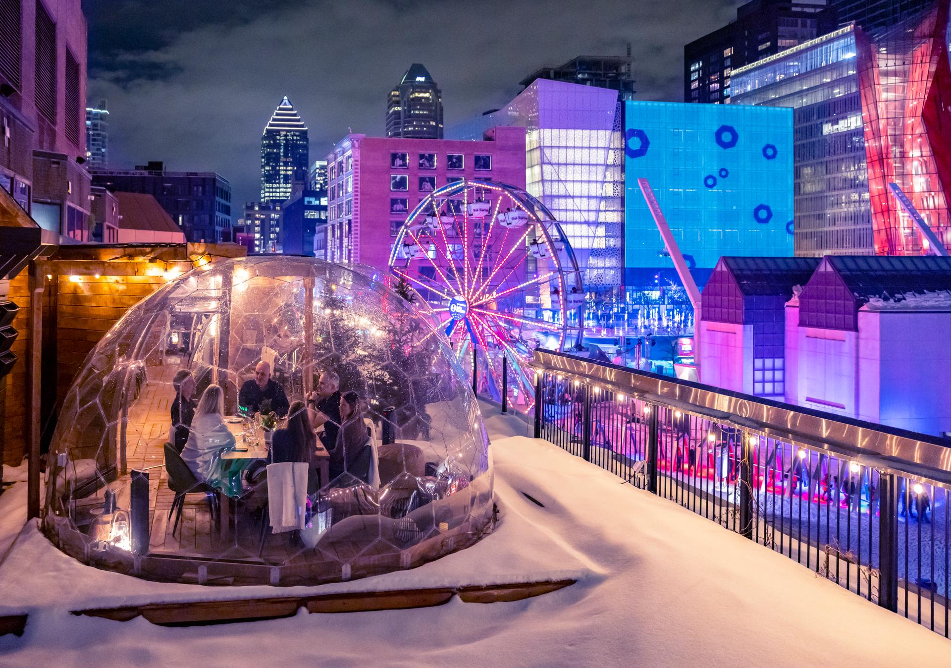 A group of people eating inside a transparent dome on a rooftop at night with downtown Montreal in the background.
