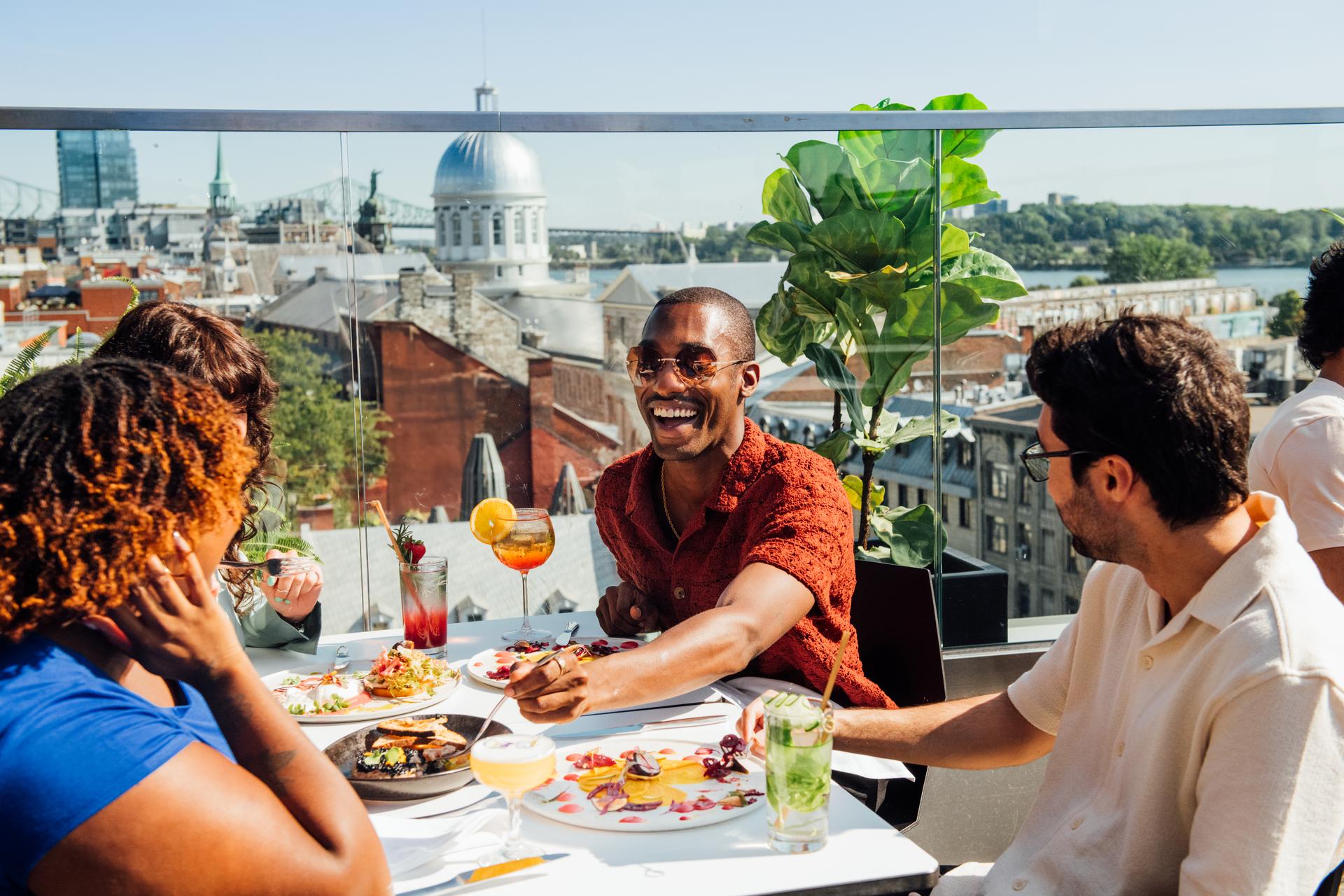 Four people enjoying a restaurant meal at a rooftop table with downtown Montreal in the background.