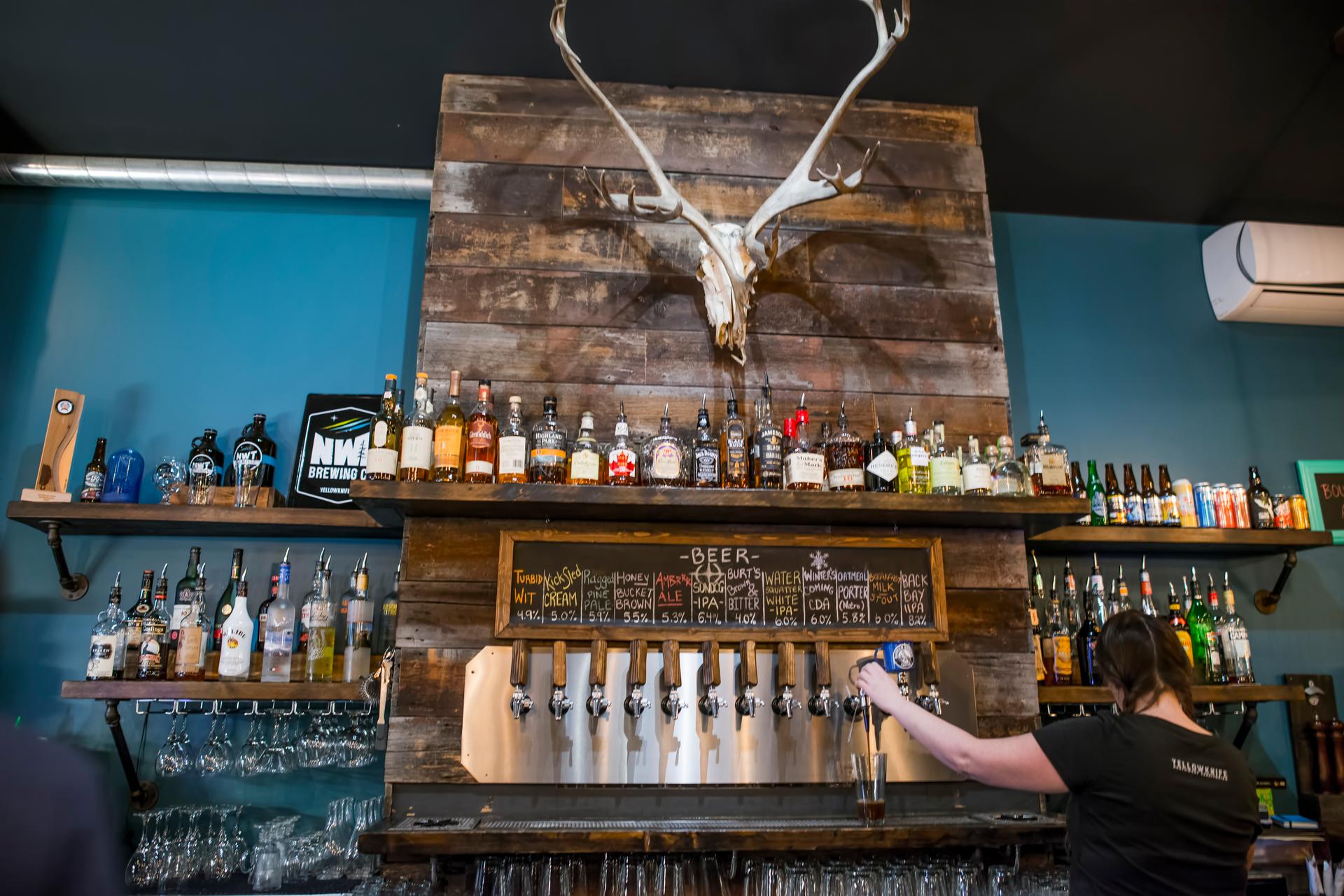 A rustic bar setup featuring a wooden backdrop with a mounted antler display, shelves stocked with various liquors and beer cans