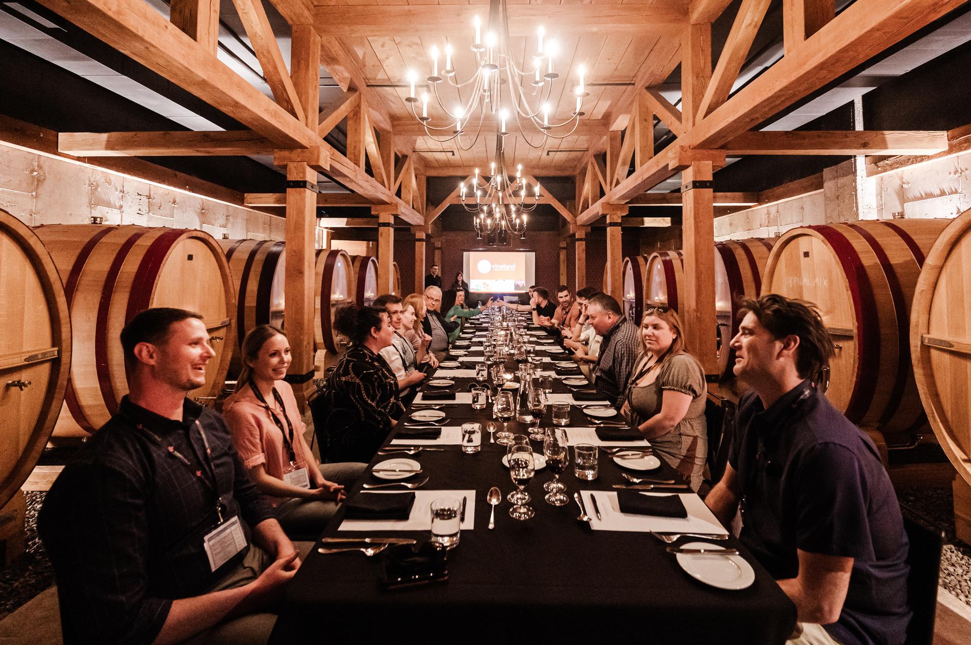 A group of people seated at a long table in a wine cellar filled with barrels.