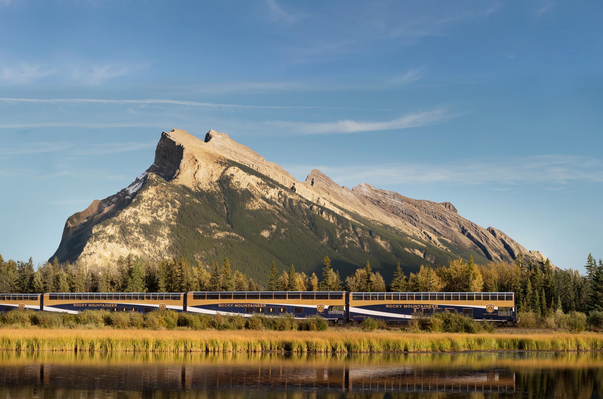 Rocky Mountaineer train passing through the Canadian Rockies with a dramatic mountain backdrop and reflection in a calm body of water