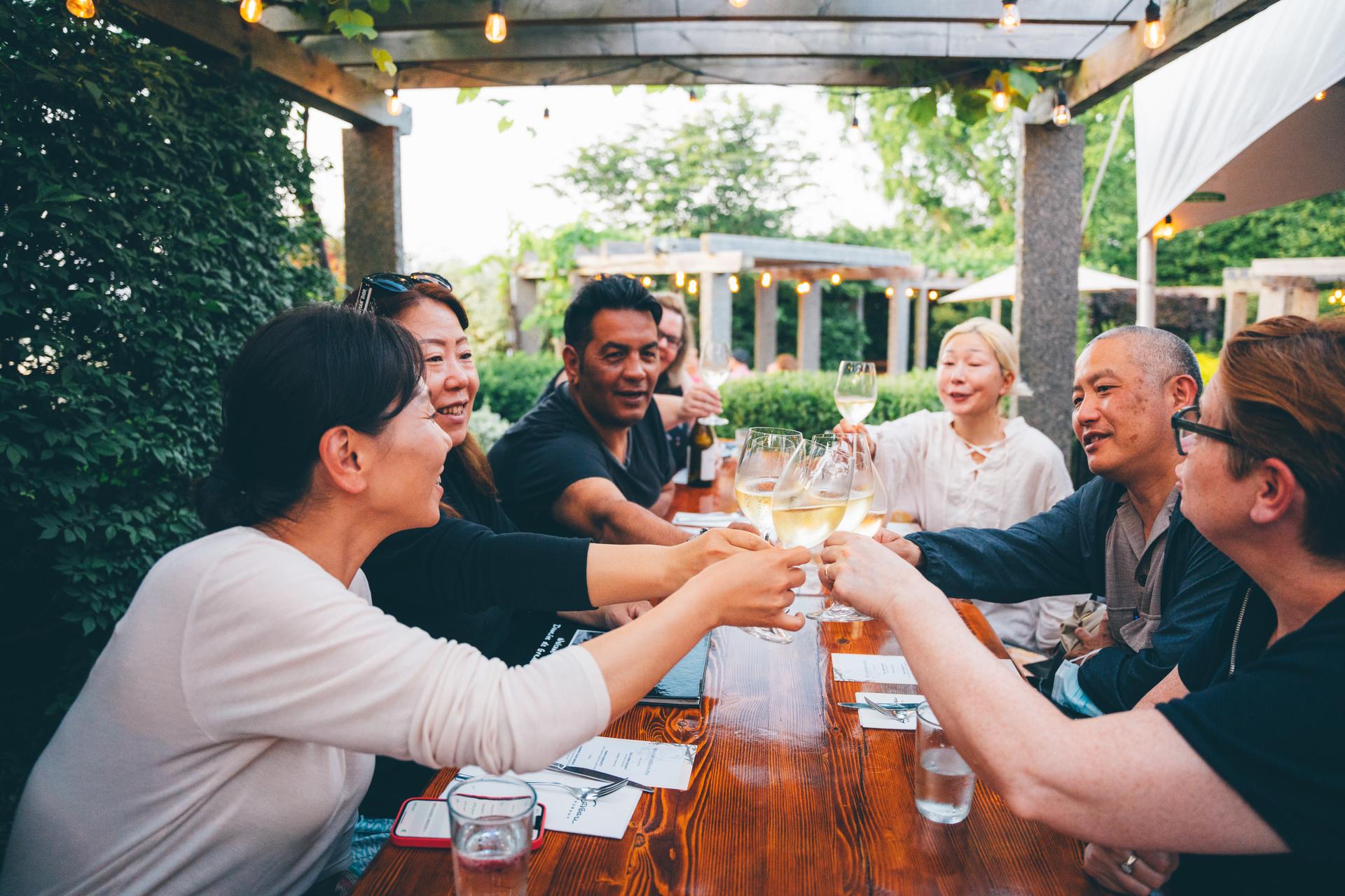 A group of people sitting at an outdoor restaurant table and clinking their glasses of wine together.
