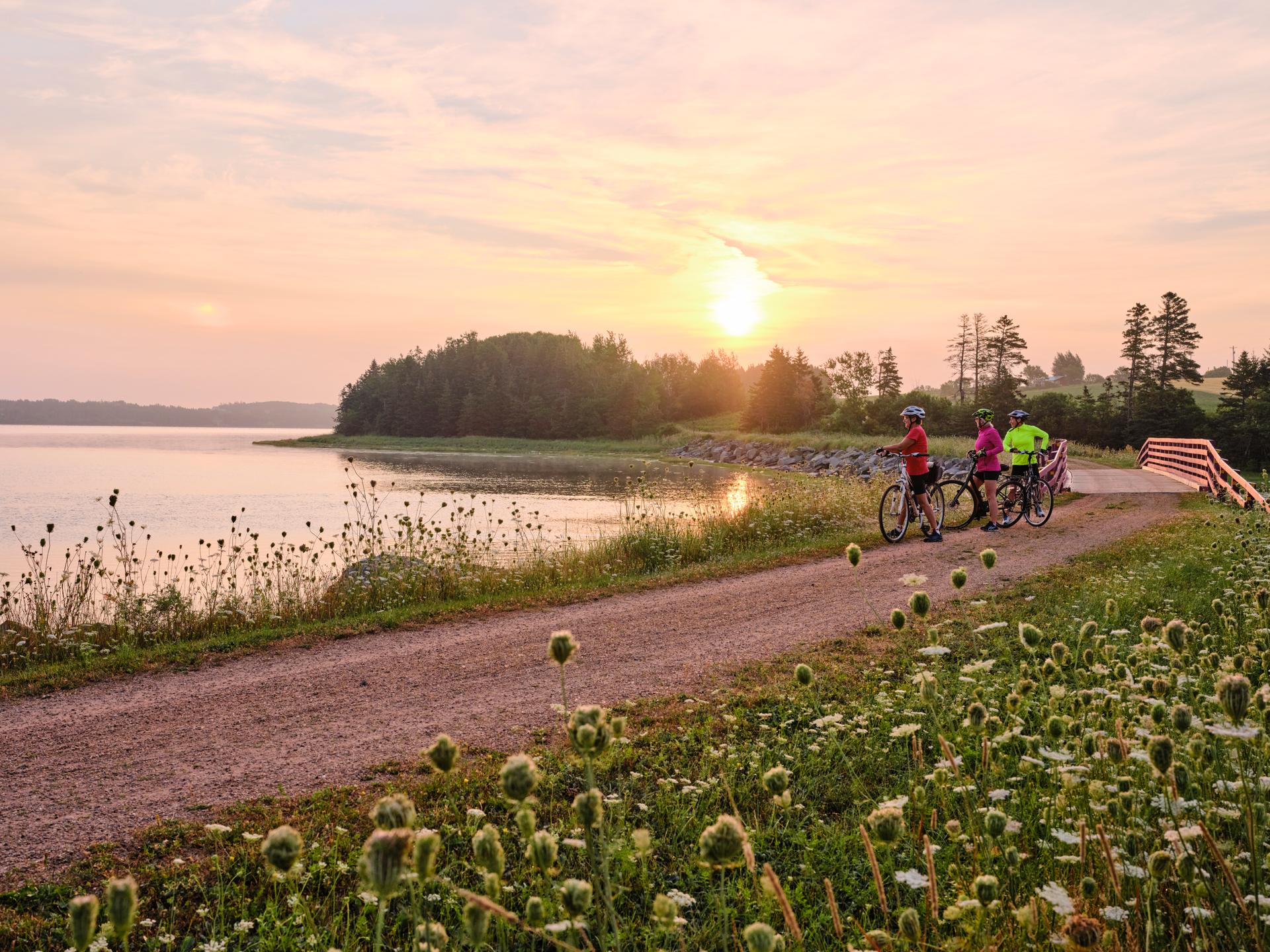 Three cyclists taking a break on a waterfront path in Prince Edward Island as the sun sets.
