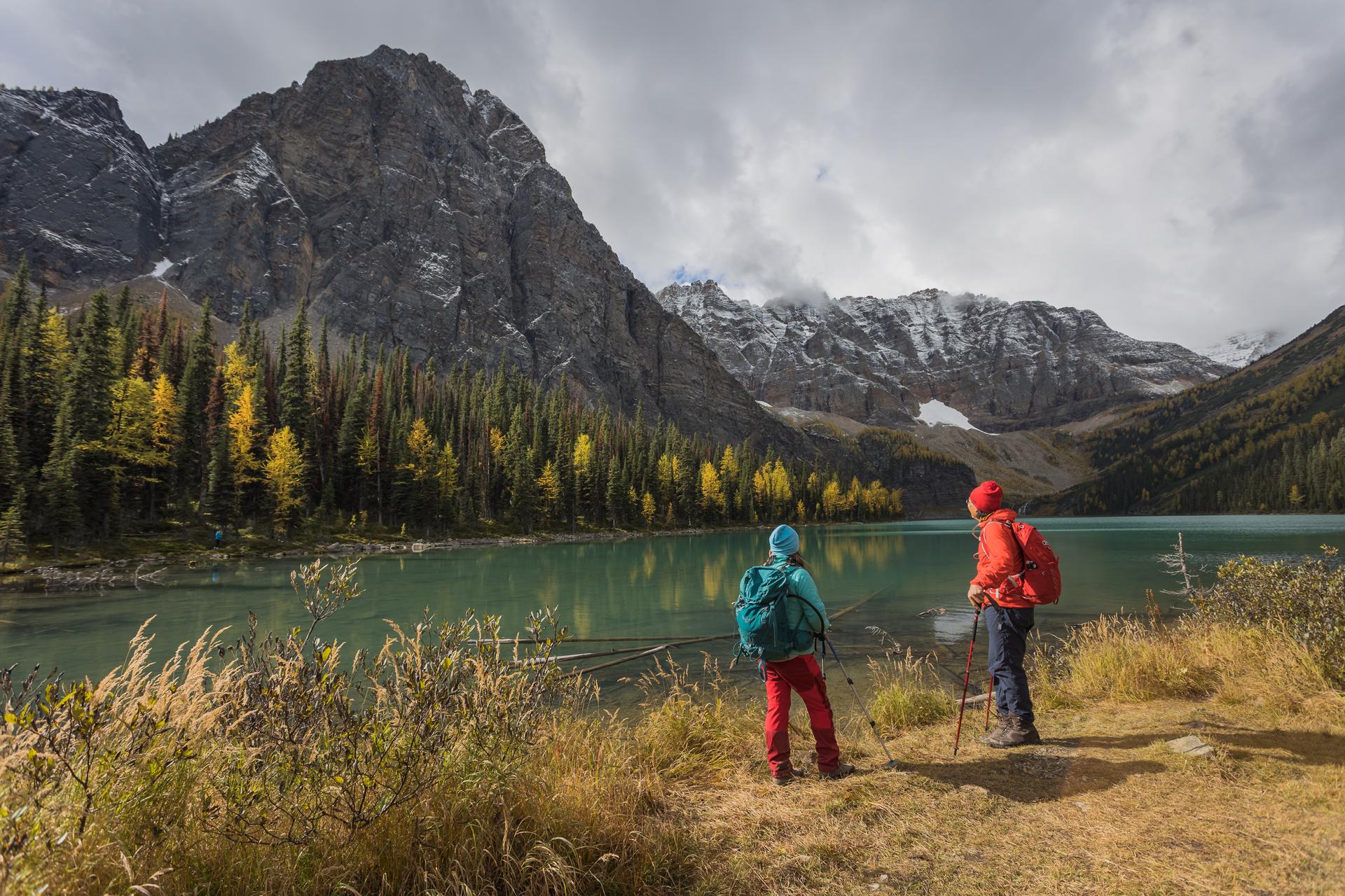 Two hikers stand by a calm mountain lake framed by trees