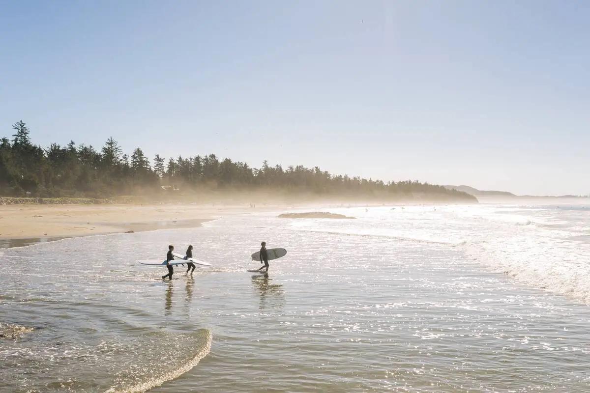 Hero image establishing Canada's beach diversity - two surfers in wetsuits walk along a misty Pacific shoreline with boards under arm, signalling that Canadian beaches offer experiences beyond tropical expectations, from surf culture to storm walks.