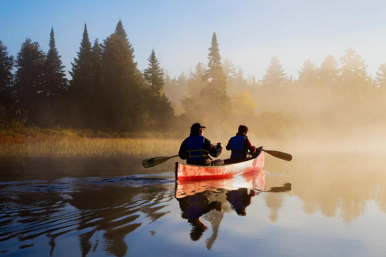 Two paddlers in a red canoe gliding across a misty Ontario lake at sunrise, surrounded by dense forest—the quintessential Canadian wilderness canoeing experience.