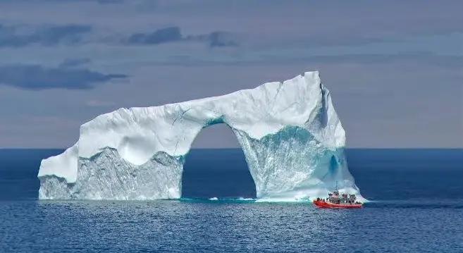 A tour boat approaches an enormous iceberg arch formation, with passengers visible on deck for scale, illustrating the office-tower size these ancient ice structures can reach.