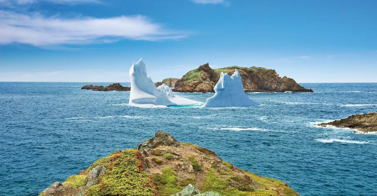 A striking aquamarine iceberg floats near the rocky Newfoundland shore with a red-roofed heritage building, highlighting how icebergs can be viewed from land at coastal villages.