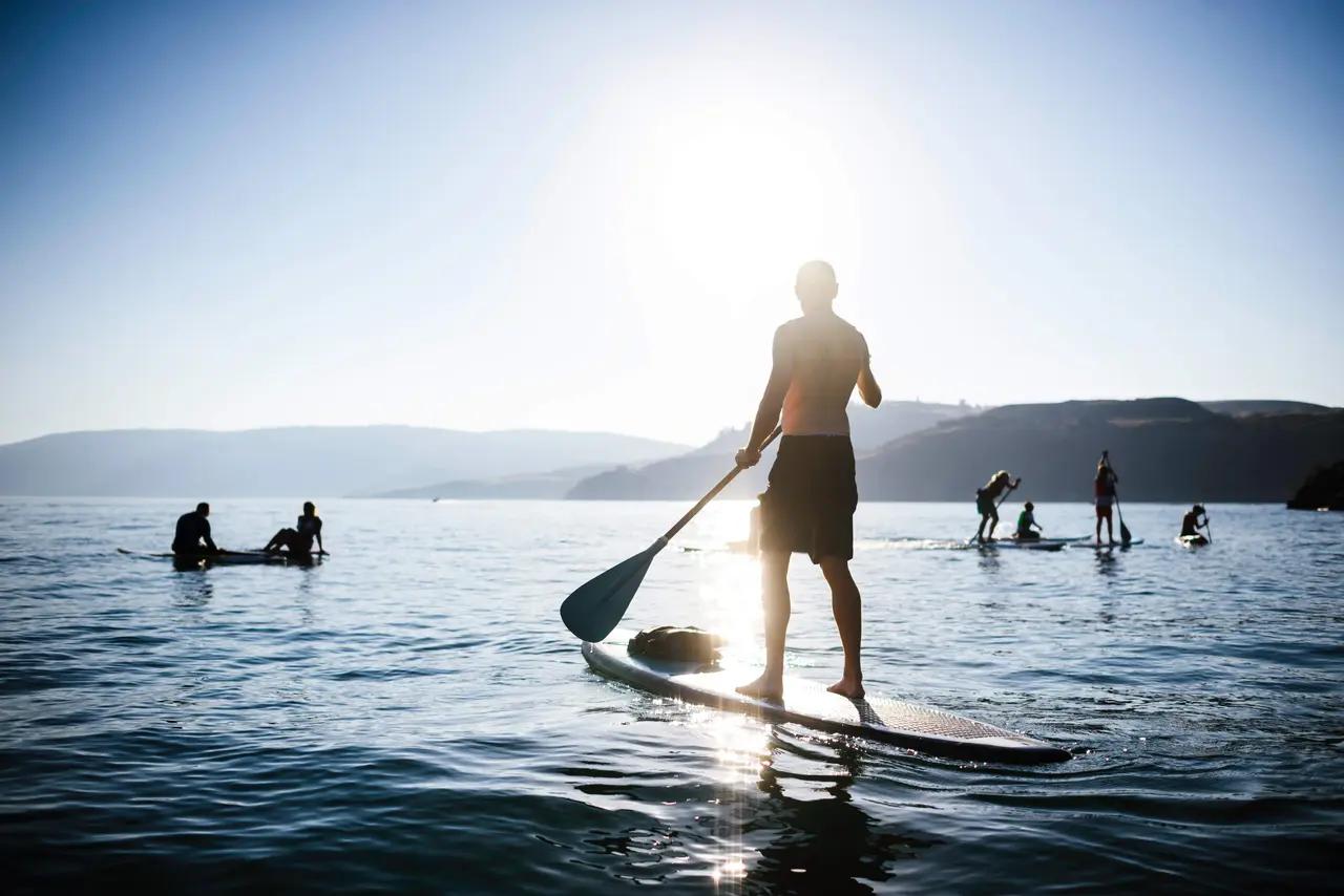 A stand-up paddleboarder glides across glassy Okanagan Lake waters at golden hour with mountains silhouetted behind, capturing the calm, warm-water conditions that make the region ideal for beginner paddlers and sunset sessions.