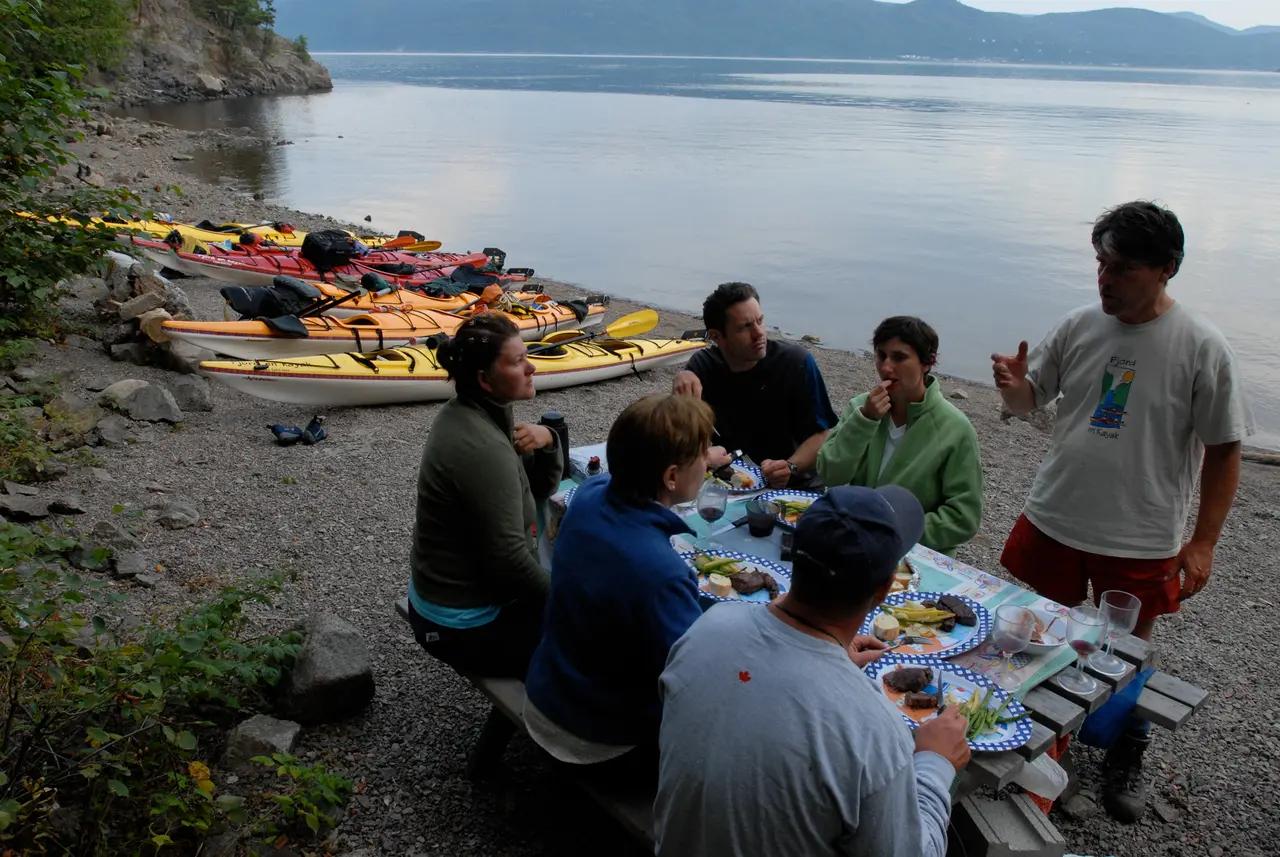 Kayakers enjoy a gourmet outdoor dinner on the shore of Saguenay Fjord during a multi-day guided adventure, with sea kayaks pulled up on the rocky beach.