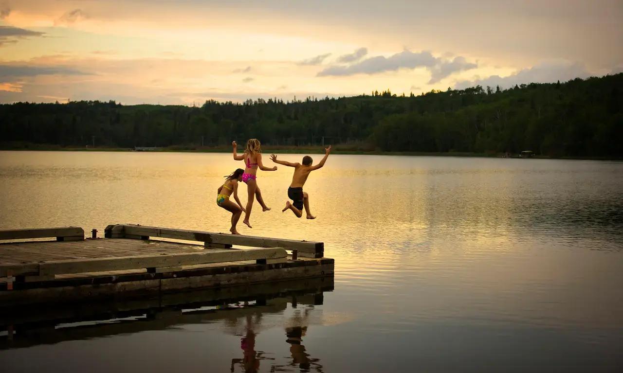 Hero image establishing Saskatchewan's lake culture - two people leap joyfully from a wooden dock into a forested lake, capturing the carefree spirit of summer holidays that defines the province's 100,000 freshwater lakes.