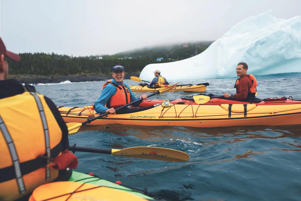 Sea kayakers paddle through calm waters near towering icebergs off the Newfoundland coast, demonstrating the intimate up-close experience available through guided kayak tours.