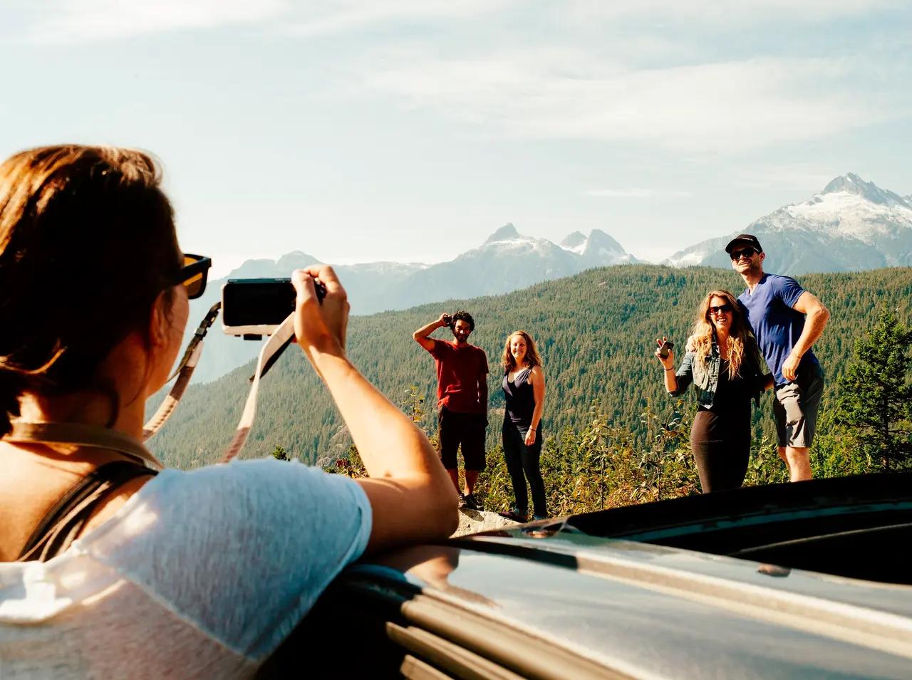 A person photographs four smiling friends posing at a mountain viewpoint along the Sea-to-Sky Highway, with dense evergreen forests, rolling green hills, and snow-capped Coast Mountain peaks stretching across the horizon behind them — capturing the rewarding stops and social experience of BC's iconic 162-kilometre drive between Vancouver and Whistler.