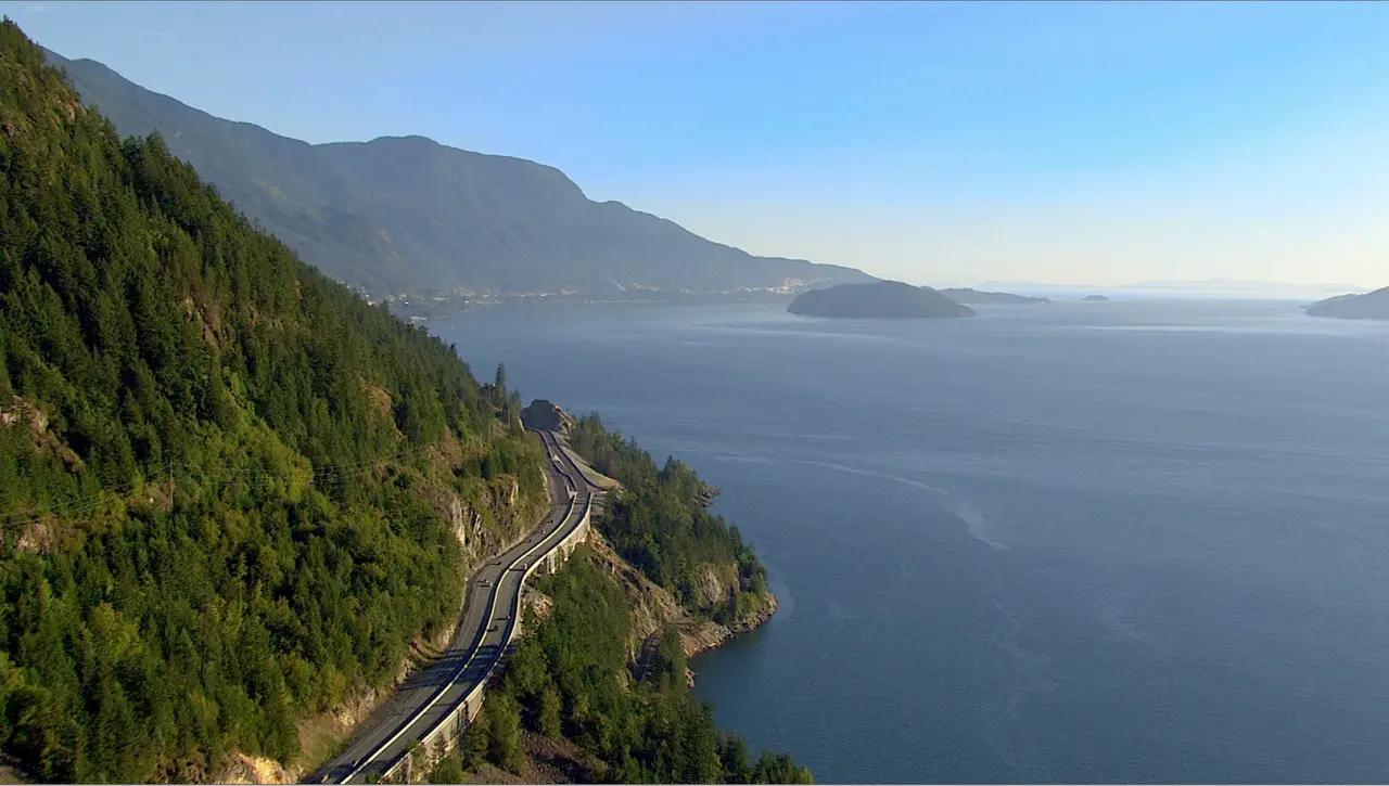 An aerial view of the Sea-to-Sky Highway carved into a steep, densely forested clifftop above the calm, dark blue waters of Howe Sound, with misty islands visible in the distance under a clear blue sky — showing the dramatic geography of BC's most iconic coastal mountain drive between Vancouver and Whistler.