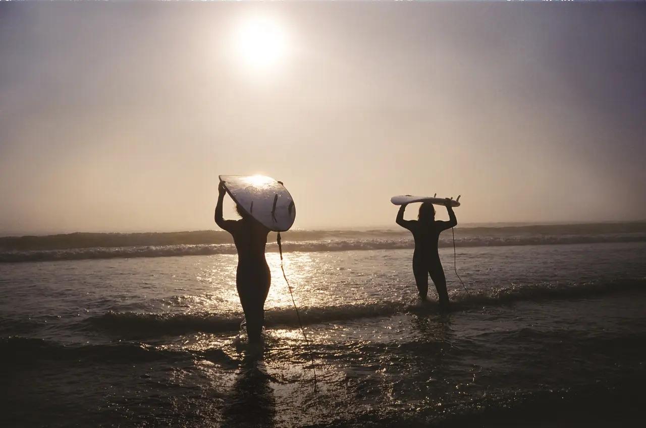 Two surfers in wetsuits silhouetted against a glowing, hazy sunset, carrying their surfboards overhead as they wade through the shallow surf on a Tofino beach — evoking the laid-back surf culture at the end of the 300-kilometre Vancouver to Tofino road trip, Canada's surf capital on the west coast of Vancouver Island.