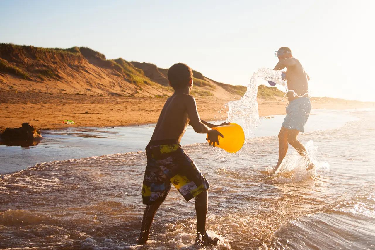 A man and child laughing and splashing each other with water on a warm, sun-drenched red sand beach, with PEI's characteristic rust-coloured sand dunes and grassy banks glowing in golden evening light behind them — Brackley Beach, a beloved family destination just 15 minutes from Charlottetown where summer days end at a classic drive-in theatre.