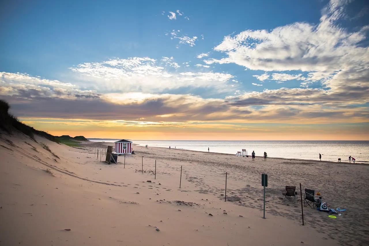 A wide, gently curving PEI beach at sunset with a small red and white striped lifeguard hut on the sand dunes, a handful of beachgoers scattered along the shoreline, and beach chairs in the foreground beneath a dramatic orange and blue evening sky — Cavendish Beach, the most popular stretch of Prince Edward Island National Park's 37-mile coastline.