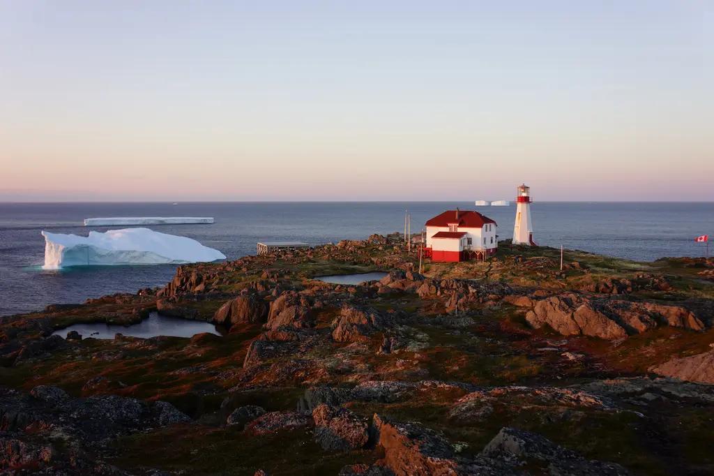 A red-roofed lighthouse keeper's house and white lighthouse tower glowing warmly at sunset on a rocky coastal headland in Newfoundland, with two large flat-topped icebergs drifting on the calm Atlantic Ocean in the background and a Canadian flag flying nearby.