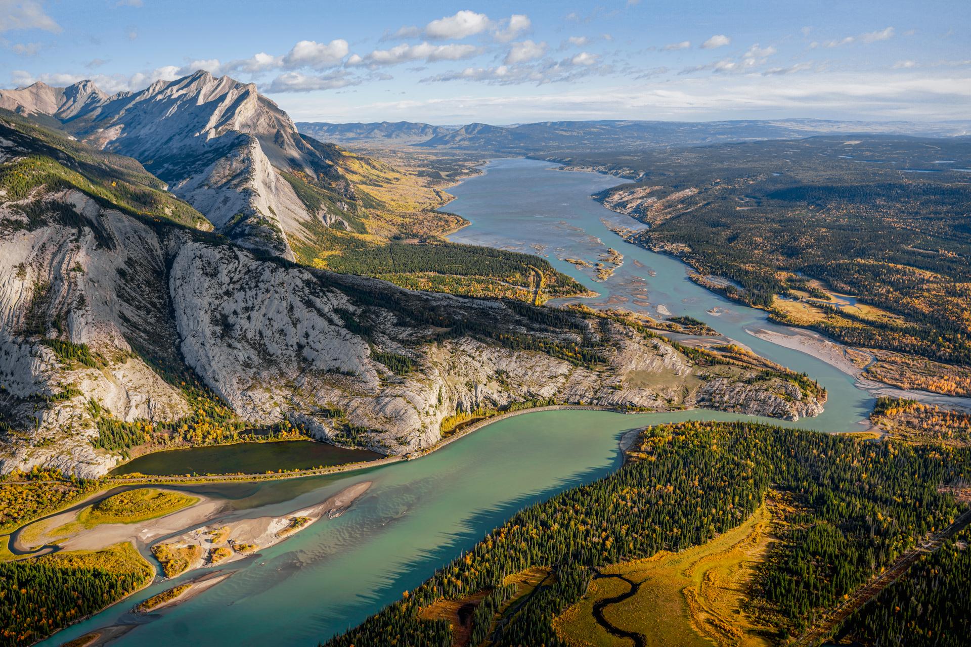 Aerial view of a glacier-fed turquoise river winding through the Canadian Rockies near Jasper, Alberta, flanked by rugged mountain cliffs and forested valleys in summer.