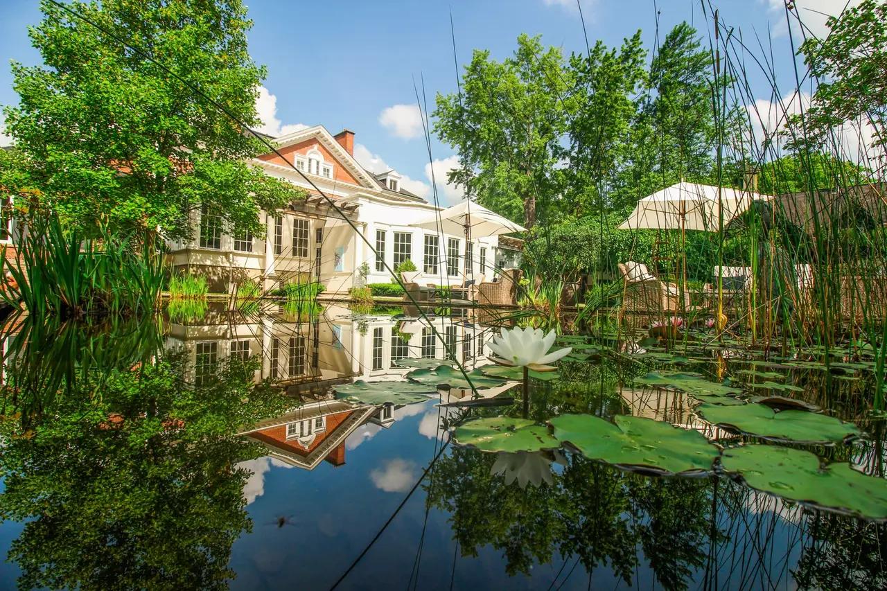 The elegant white Georgian facade of Langdon Hall Country House Hotel and Spa perfectly reflected in a tranquil lily pond, with white water lilies blooming, tall reeds, lush mature trees, and outdoor terrace umbrellas visible on a sunny summer day — a Relais & Châteaux manor in Cambridge, Ontario offering manicured gardens, spa treatments, fireside dining, and the kind of romantic, unhurried setting designed for complete relaxation.