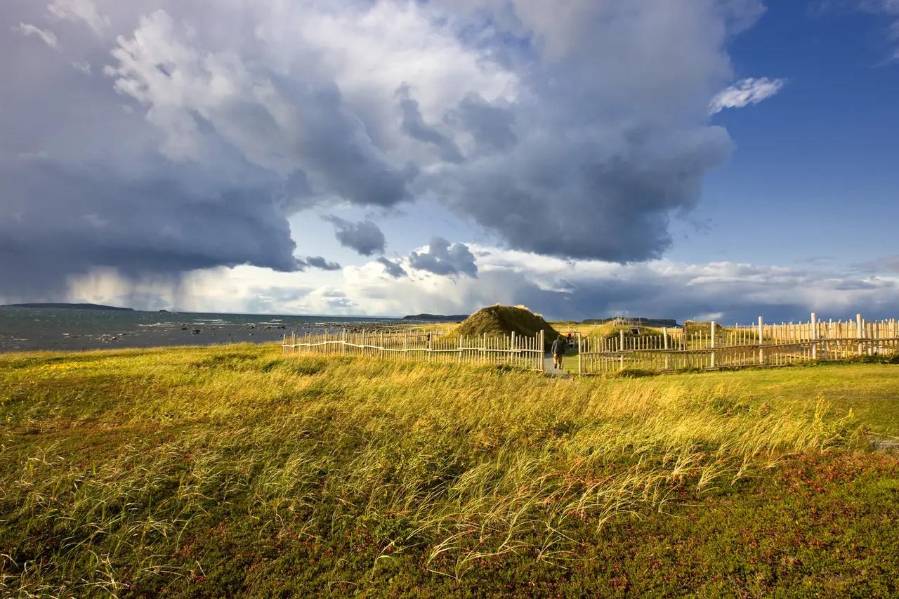 A visitor walks a wooden pathway toward a reconstructed Norse sod longhouse at L'Anse aux Meadows National Historic Site, Newfoundland, with wind-swept coastal grasses in the foreground, a dramatic storm rolling in over the ocean, and wooden palisade fencing marking North America's only confirmed Viking settlement.
