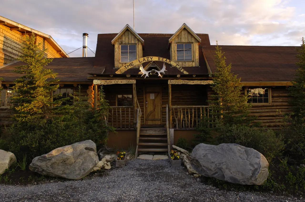 The rustic log exterior of The Lazy Bear Cafe at Lazy Bear Lodge in Churchill, Manitoba at golden hour, with a hand-carved wooden sign, moose antler decoration above the entrance, and menu boards advertising musk ox, Arctic char, and caribou — the National Geographic Traveler top-rated lodge built from reclaimed heritage materials that serves as a cosy home base for polar bear tours and beluga whale watching on Hudson Bay.