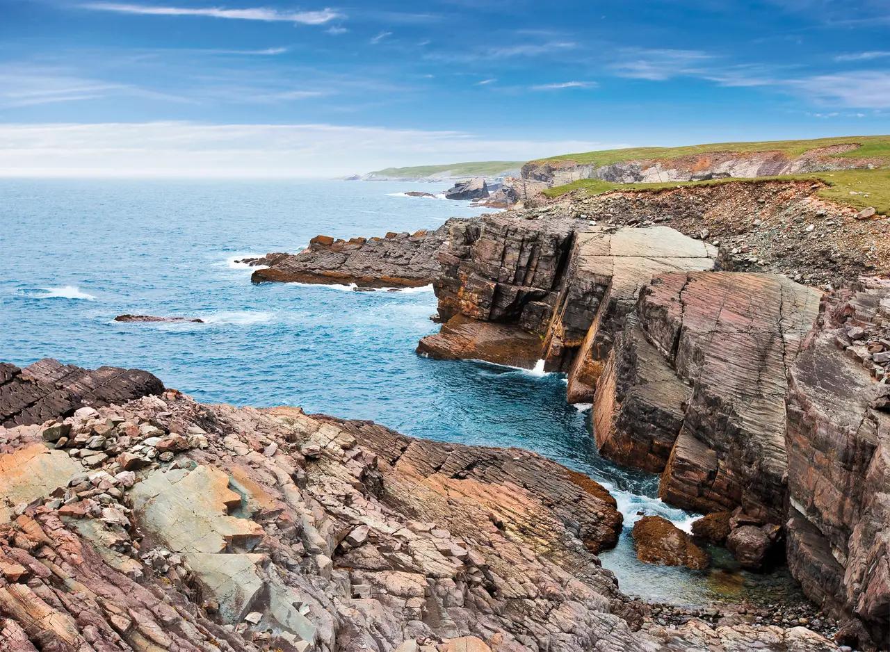 Dramatically layered and fractured ancient rock formations along the Newfoundland coastline at Mistaken Point Ecological Reserve, where vivid rust, grey, and copper-toned sedimentary cliffs drop into narrow sea channels of brilliant blue Atlantic water under a bright sky.