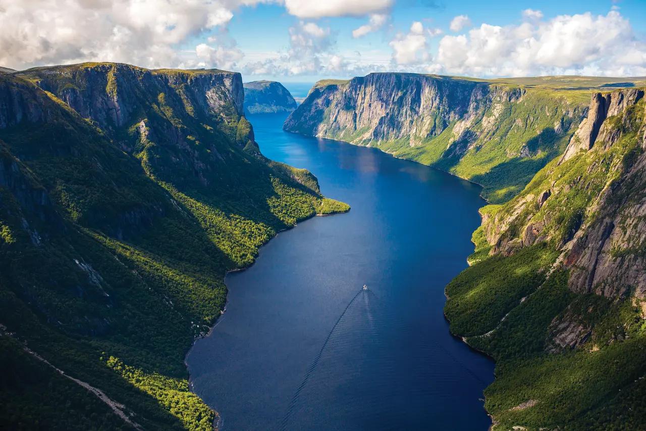 An aerial view looking down the length of Western Brook Pond fjord in Gros Morne National Park, Newfoundland, with a tiny tour boat trailing a white wake through deep blue water flanked by towering green-forested cliff walls stretching into the distance.