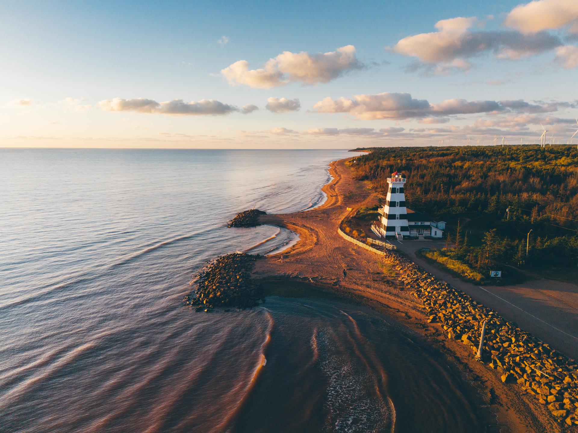 Aerial sunset view of West Point Lighthouse on a narrow red sand spit along the coastline of Prince Edward Island, Canada, with calm ocean waters, rocky breakwaters, and forested shoreline stretching into the distance