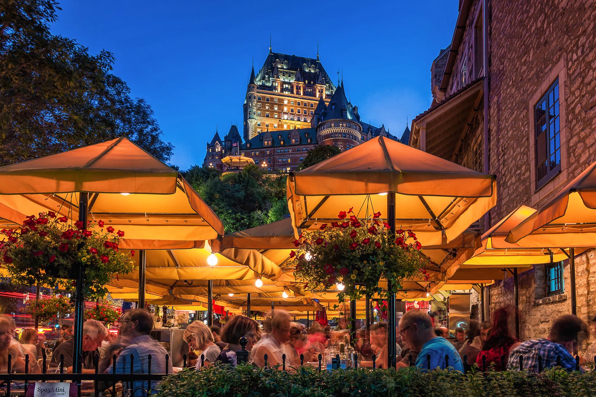 Outdoor dining terrace in Old Quebec City at night, with guests seated under lit amber umbrellas along a historic cobblestone street, and the illuminated Château Frontenac hotel visible above.