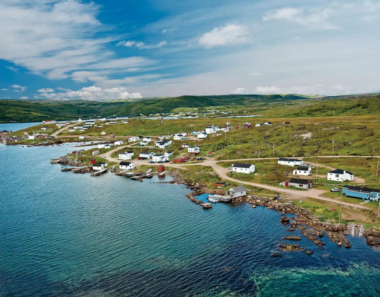 An aerial view of the small coastal community of Red Bay, Labrador, with white and red clapboard fishing buildings and wooden docks lining a rocky shoreline, rolling green hills stretching inland, and the calm blue waters of the Strait of Belle Isle surrounding the historic Basque whaling harbour.