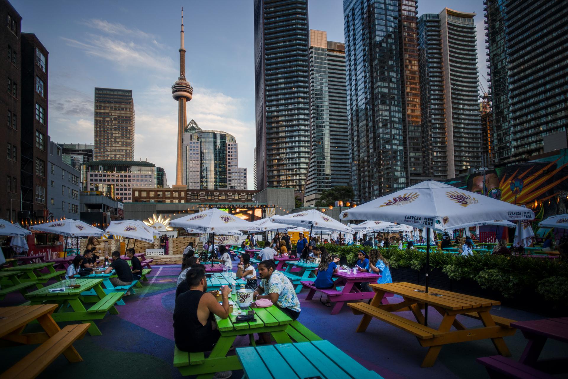 Vibrant outdoor patio with colourful picnic tables and market umbrellas filled with summer diners, set against Toronto's downtown skyline and CN Tower at dusk.