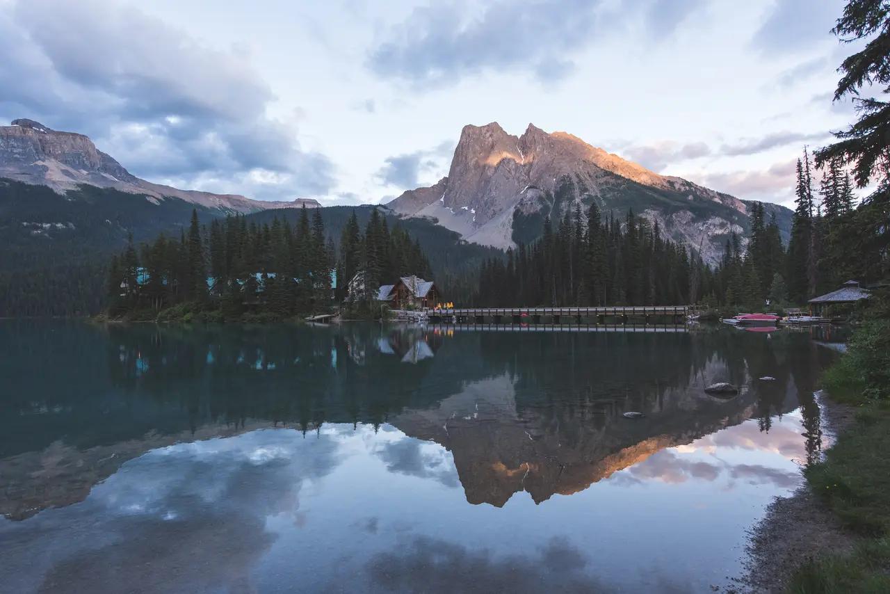 A glassy mountain lake perfectly reflecting a rustic wooden lodge, dense evergreen forest, and a towering rocky peak at dusk in Yoho National Park, British Columbia — the serene, unhurried beauty that defines Canada as a destination for rest and rejuvenation.