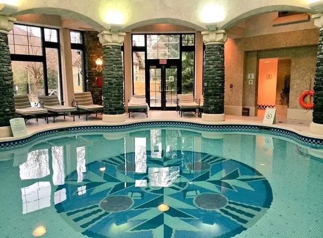 The elegant indoor mineral pool at Fairmont Spa, featuring a decorative mosaic tile floor, stone columns and lounge chairs surrounding the calm turquoise water.