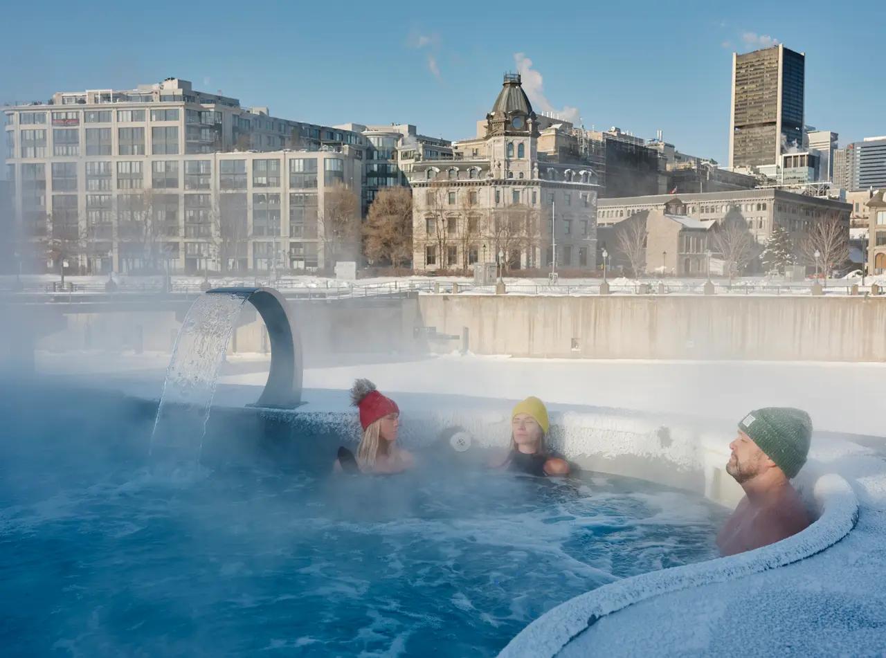 Three people relax in a steaming outdoor thermal pool at Bota Bota, spa-sur-l’eau in winter, with the historic buildings of the Old Port of Montréal visible in the snowy background.