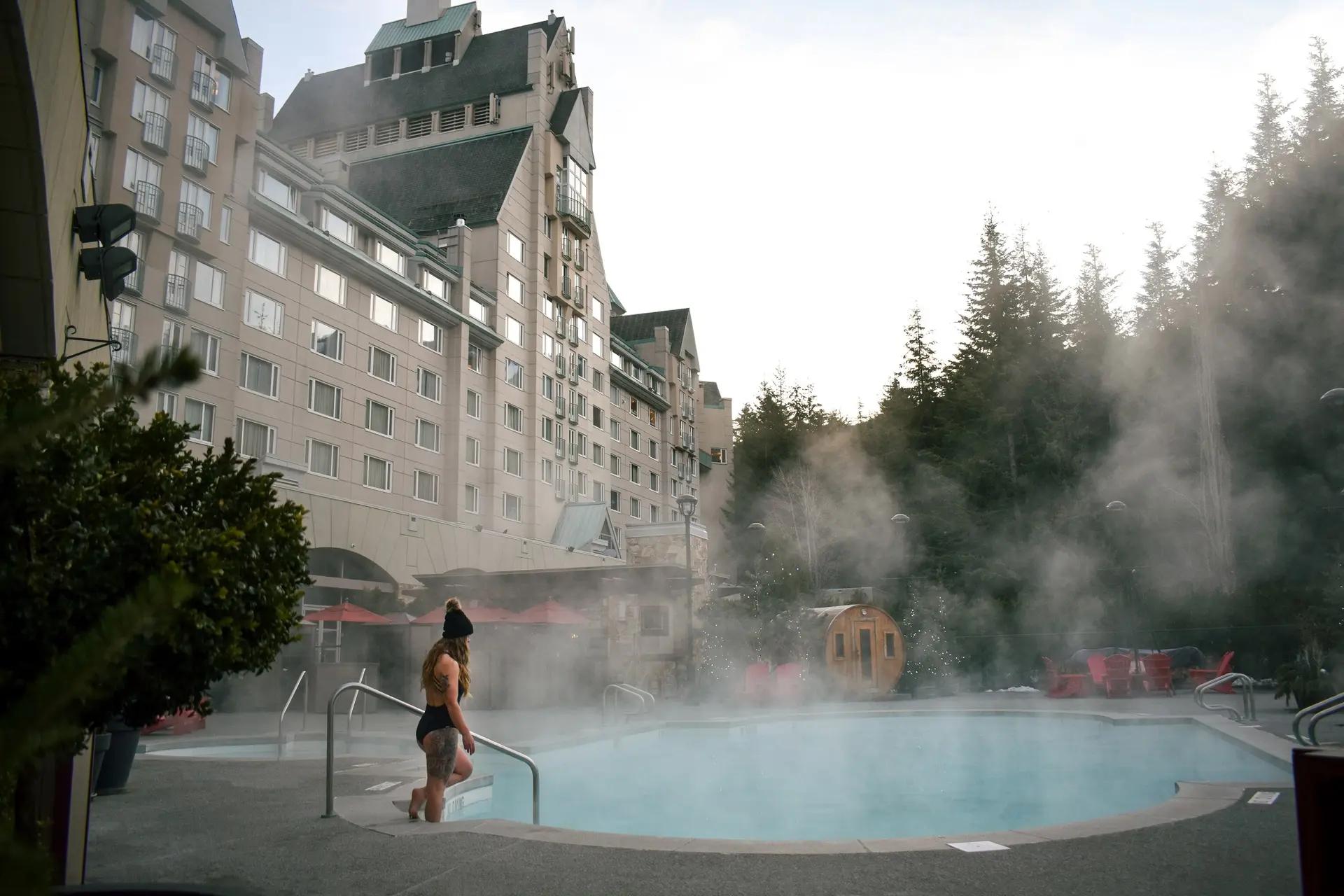 Woman stepping into the steaming outdoor pool at Fairmont Chateau Whistler with barrel sauna and evergreen forest in winter.