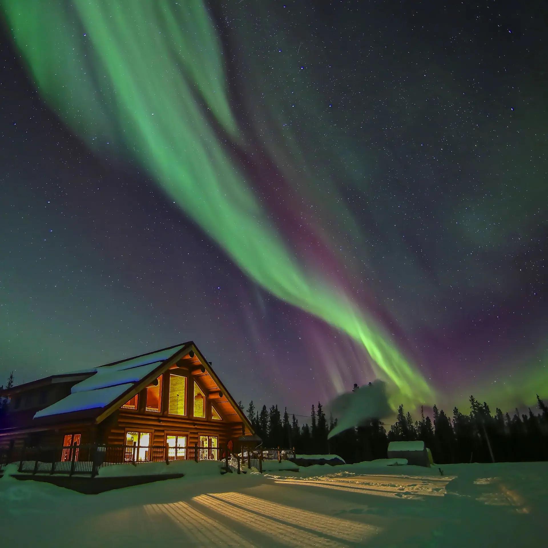 Brilliant green and purple aurora borealis streak across the night sky above the log cabin lodge at Northern Lights Resort and Spa.