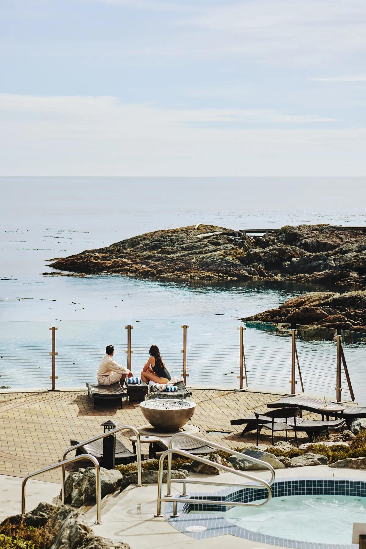 A couple sits on a waterfront deck at the Boathouse Spa at Oak Bay Beach Hotel, overlooking the rocky coastline and calm waters of the Strait of Juan de Fuca.