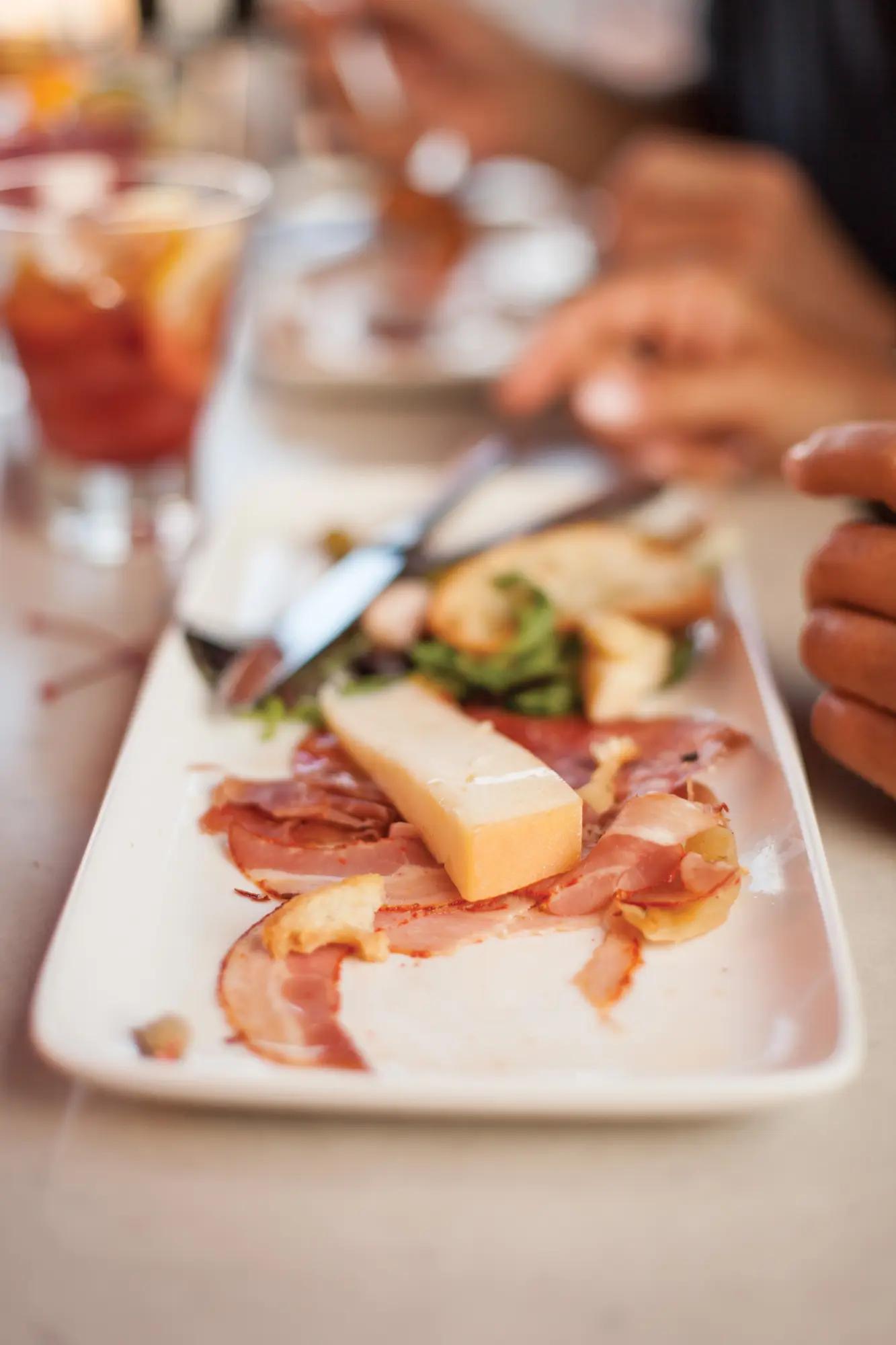 Close-up of cured prosciutto and aged cheese on a white plate at an Ottawa restaurant with bread and greens in the background