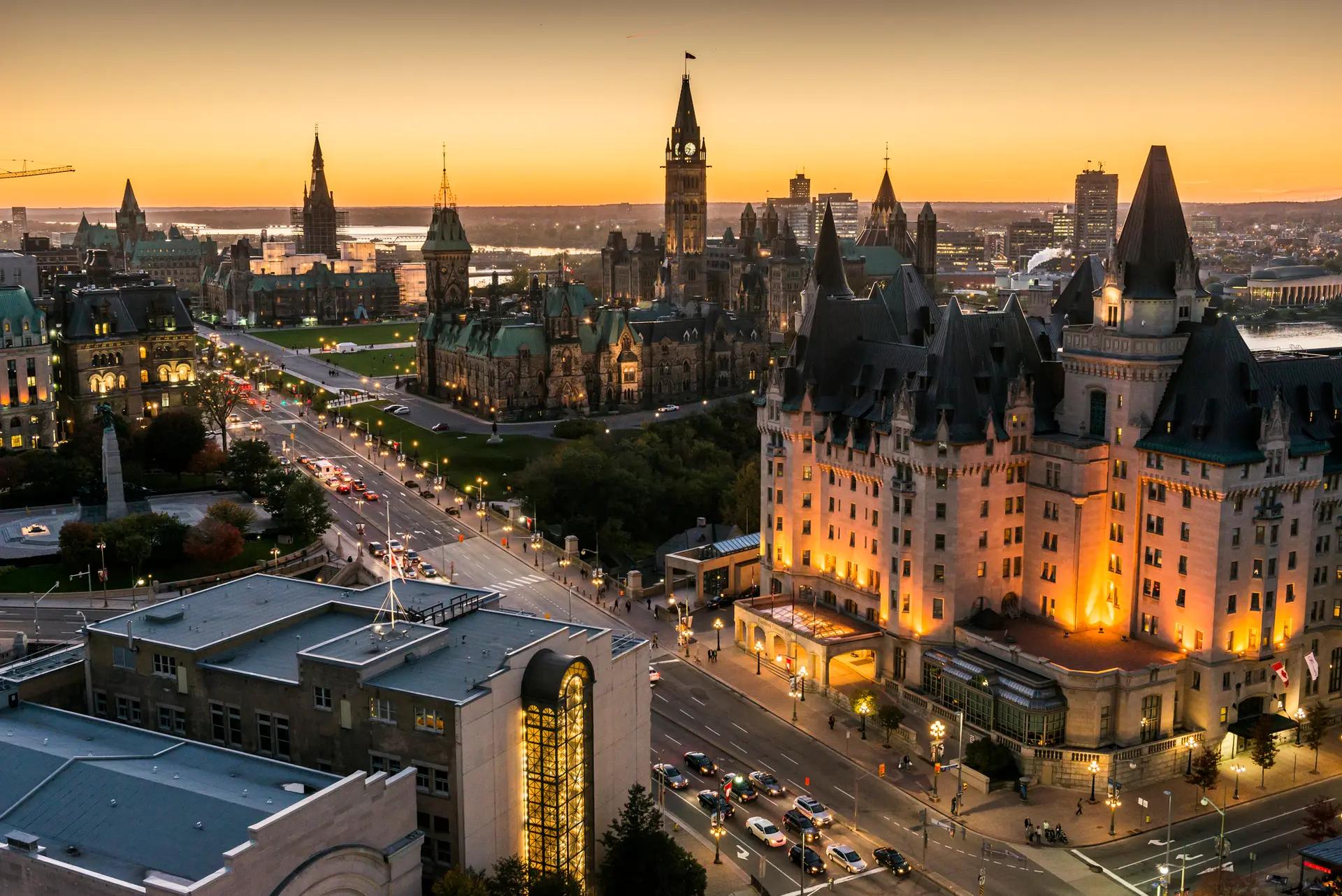 Aerial view of Ottawa's illuminated downtown skyline at sunset with Parliament Hill and the Château Laurier glowing against an orange sunset
