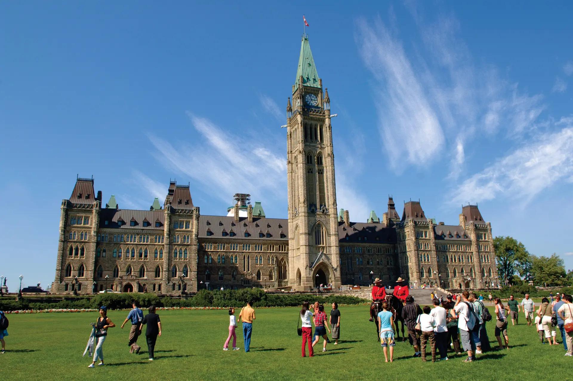 Visitors and mounted police officers in red uniforms gather on the lawn of Parliament Hill under a bright blue summer sky