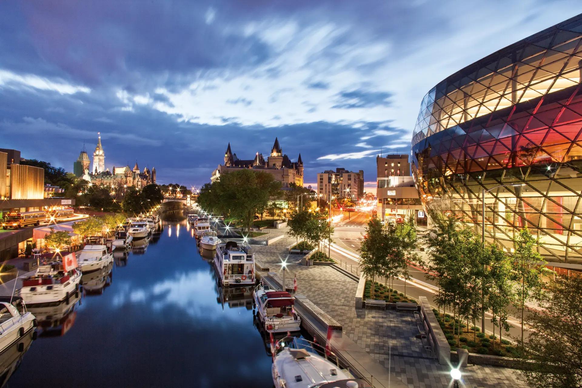Boats docked along Ottawa's Rideau Canal at dusk with Parliament Hill's illuminated Gothic towers in the background
